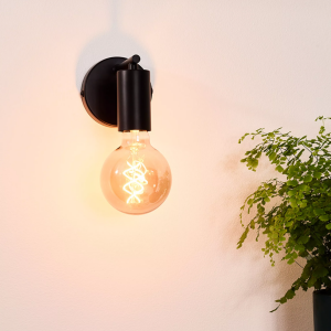 Zwarte wandlamp met zichtbare gloeilamp en warm licht naast groene plant