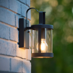 Zwarte cilindervormige buiten wandlamp met helder glas aan gevel bij schemering