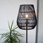 Zwarte rotan vloerlamp met opengewerkte kap en warm licht in moderne hoek
