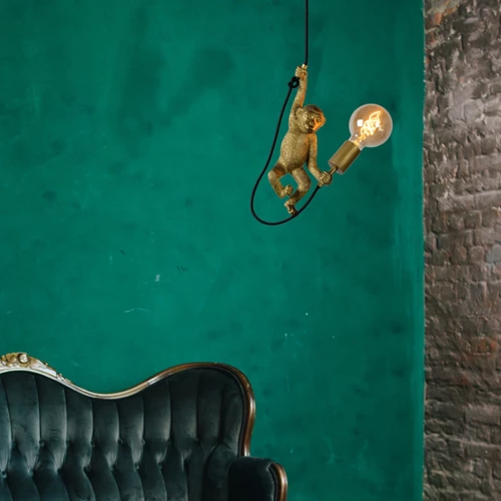 Gouden hanglamp in apenvorm met zichtbare gloeilamp boven groene muur en bank