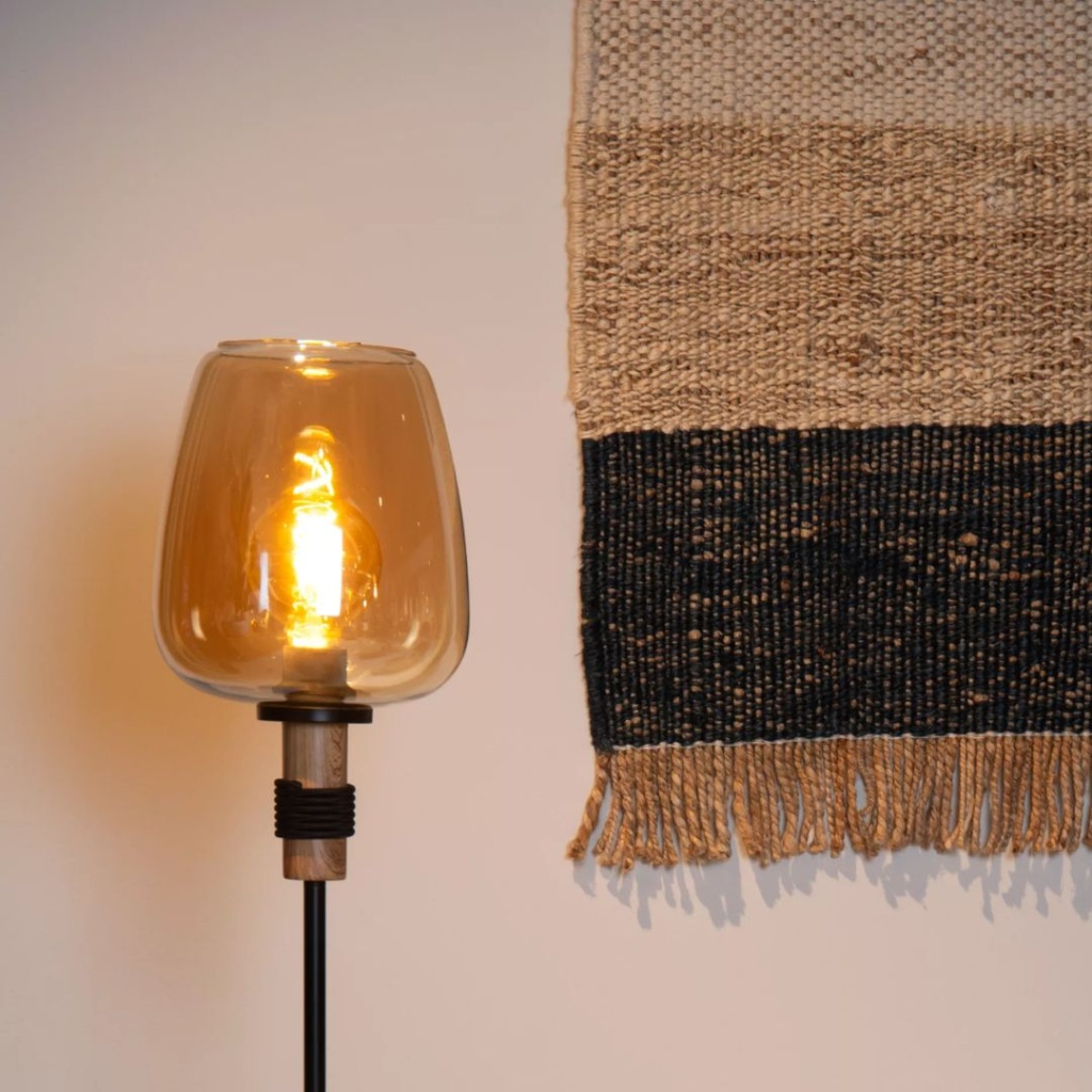 Amberkleurige vloerlamp met glazen kap en warm licht naast wandkleed